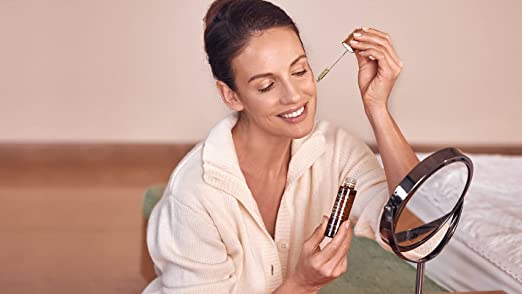 Woman applying Ahava facial serum with dropper, using mirror, relaxing at home, promoting natural beauty, cosmetic treatment, anti-aging products, health and wellness, Dead Sea minerals, skincare routine, beauty care products.