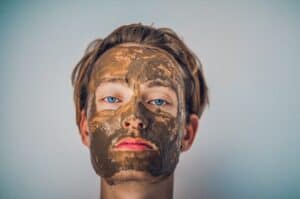 Young man with blue eyes applying mineral-rich Dead Sea mud facial mask for skin care and rejuvenation.
