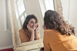 Beautiful woman smiling at her reflection in a vintage mirror, showcasing radiant skin, in a cozy natural light setting, emphasizing Dead Sea cosmetics benefits.