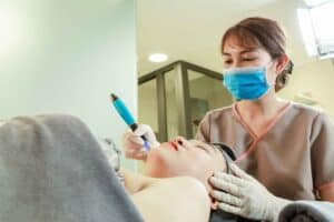 Friendly skincare technician applying Dead Sea mud mask and facial treatment to a young woman in a beauty clinic setting.