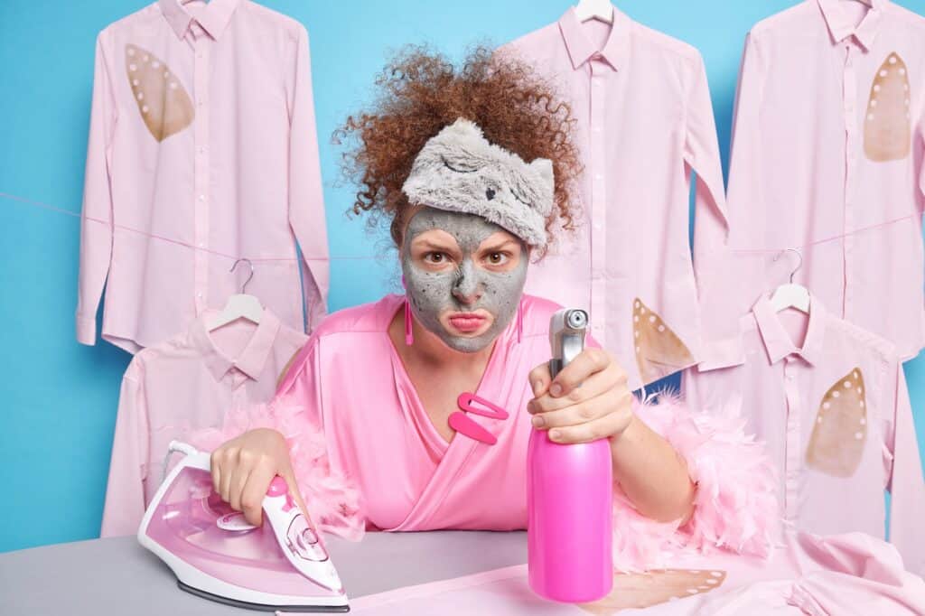 Curly-haired woman in facial clay mask doing chores surrounded by clothing, highlighting chemical exposure and skin stress